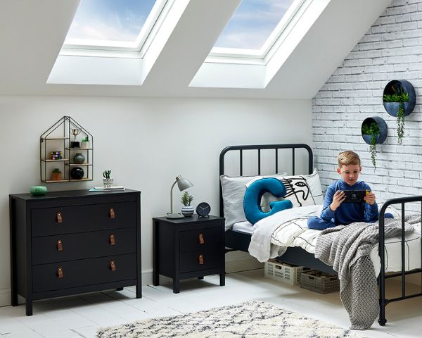 boy sat on black metal bed next to black chest of drawers with shelving above.