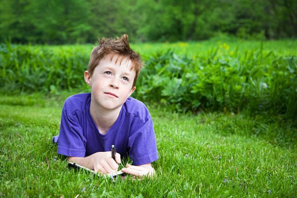 Boy lying in grass outside, writing in a notebook or journal.